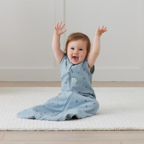 Baby in a blue sleeping bag with a small logo, sitting on a light-colored floor.