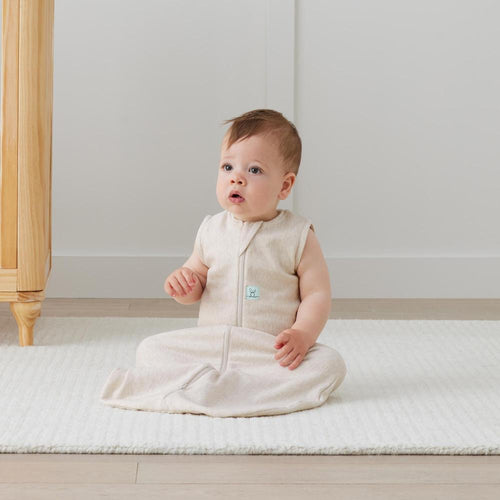 Baby sitting on a white mattress wearing a beige sleeveless outfit with a small logo.