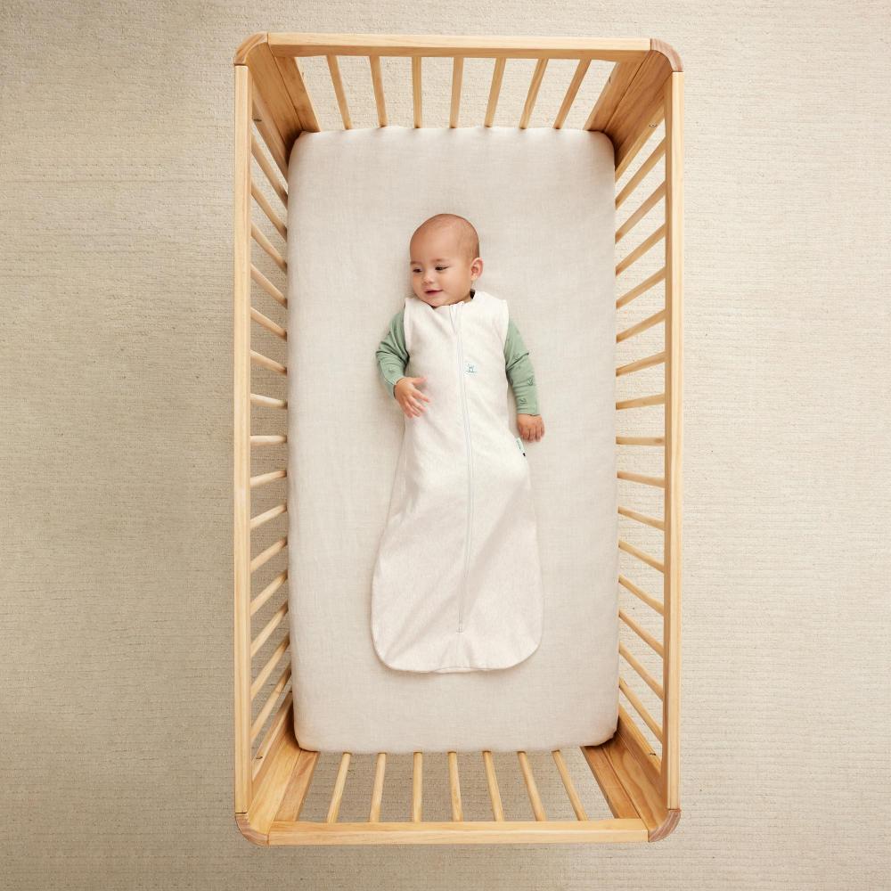 Baby in a white sleeping bag lying in a wooden crib against a beige wall.