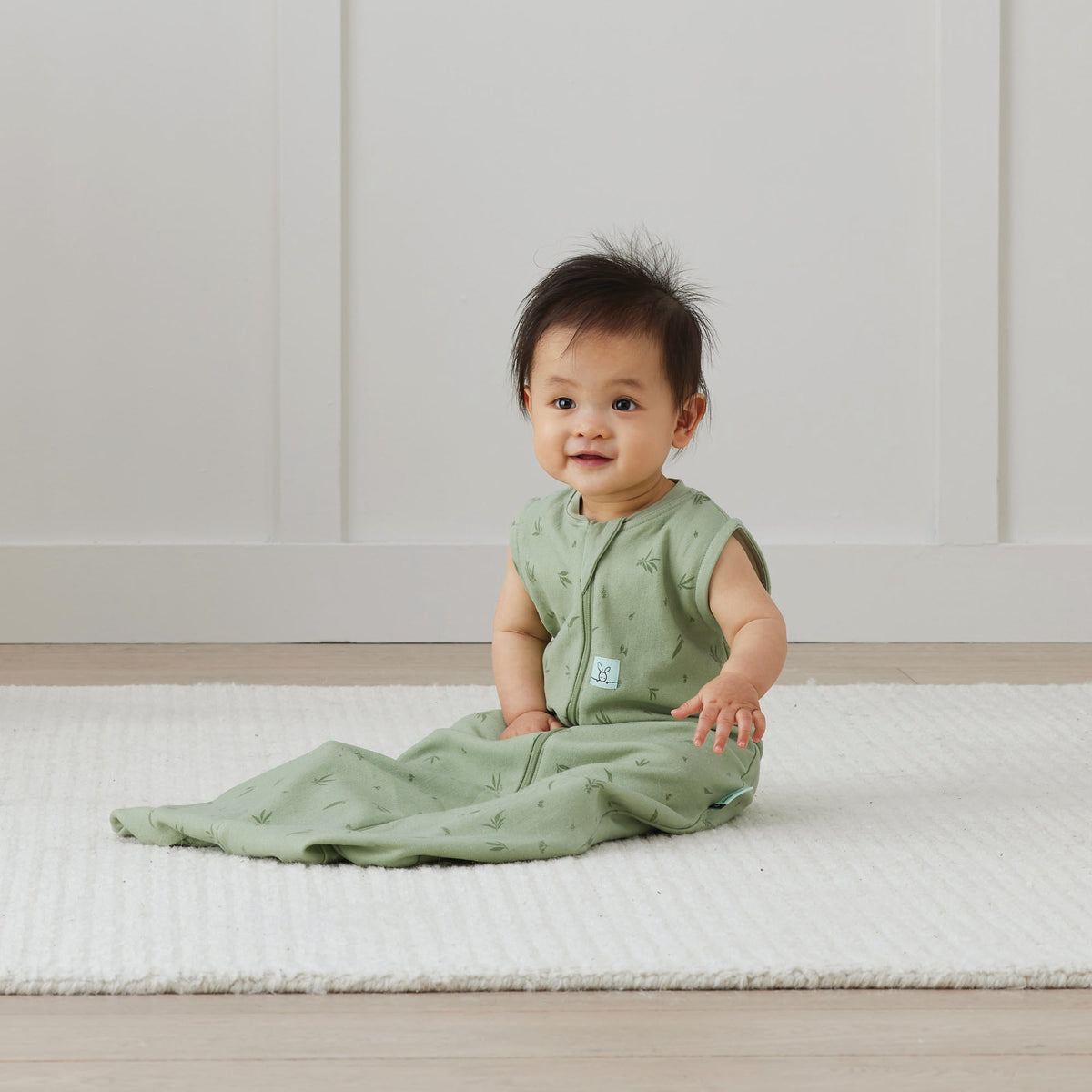 Baby in a green outfit sitting on a white mat with a white wall background