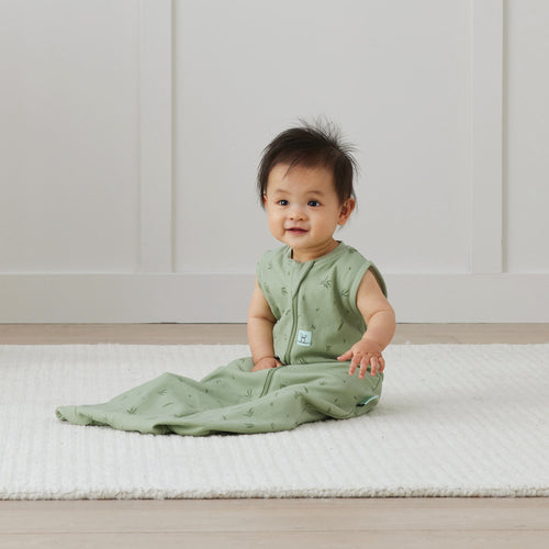 Baby in a green sleeping bag sitting on a white mat with a white wall background