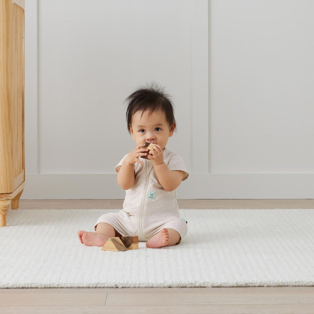 Baby in a beige romper sitting on a white mat in a minimalistic room