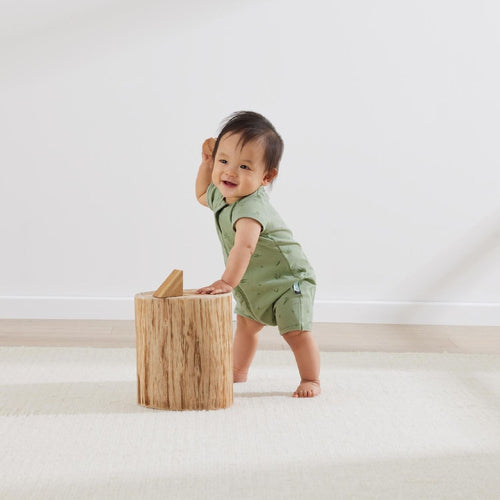 Baby in a green outfit standing next to a wooden stool on a white floor.