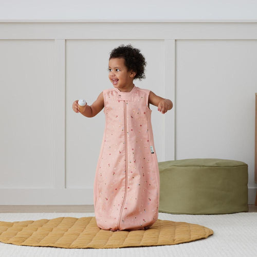 Child wearing a pink sleeveless sleep sack with a pattern, standing on a yellow mat in a room.