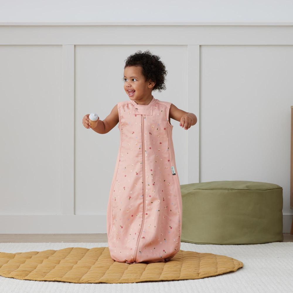 Child wearing a pink sleeping bag with a pattern, standing on a yellow mat in a room.