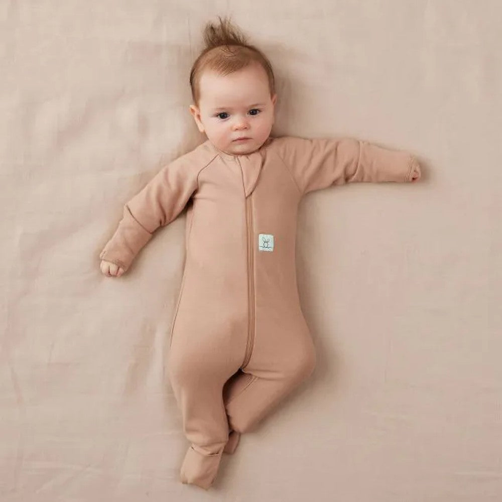 Baby lying on a beige blanket wearing a brown onesie with a visible brand tag.