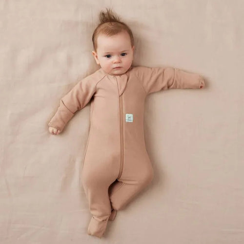 Baby lying on a beige blanket wearing a brown onesie with a visible brand tag.