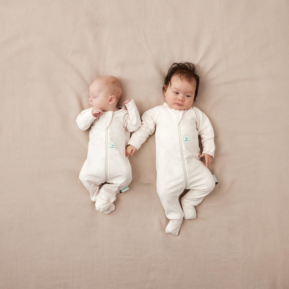 Two babies wearing beige onesies lying on a beige surface.