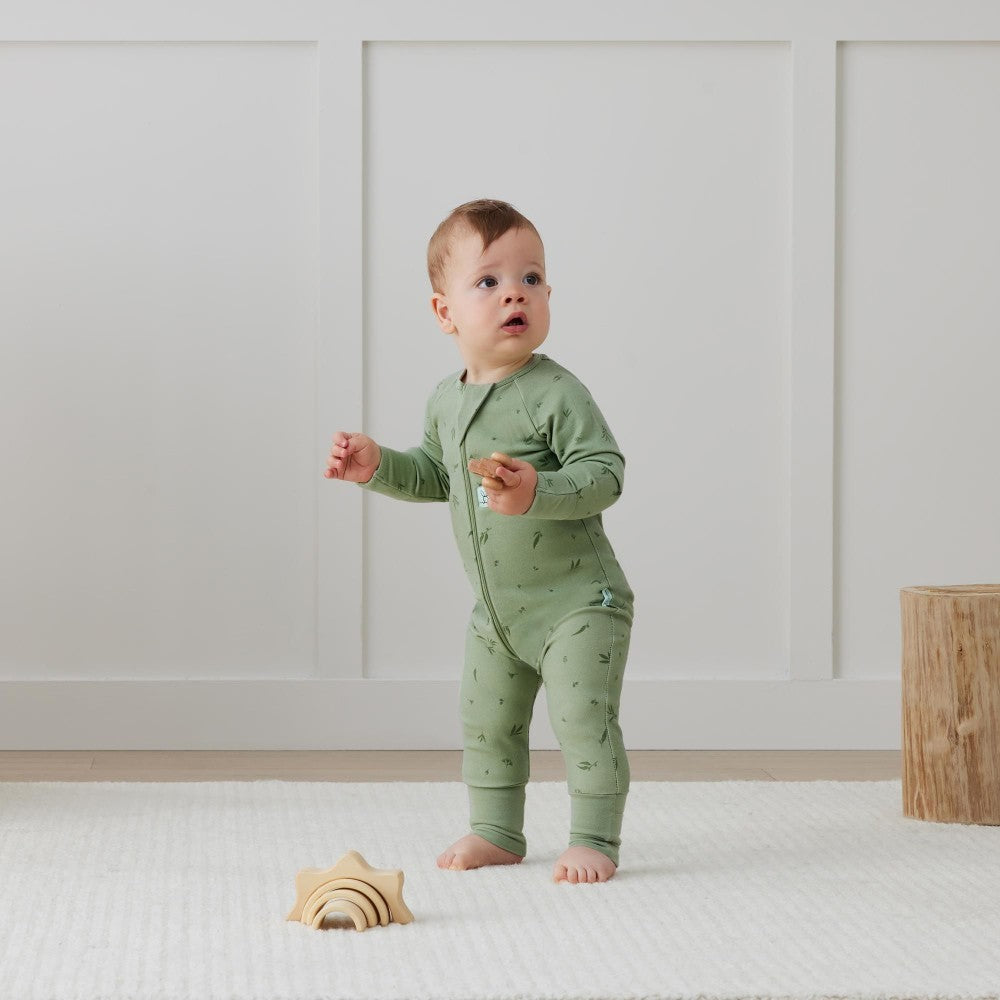 Baby in a green onesie standing in a room with a wooden toy.