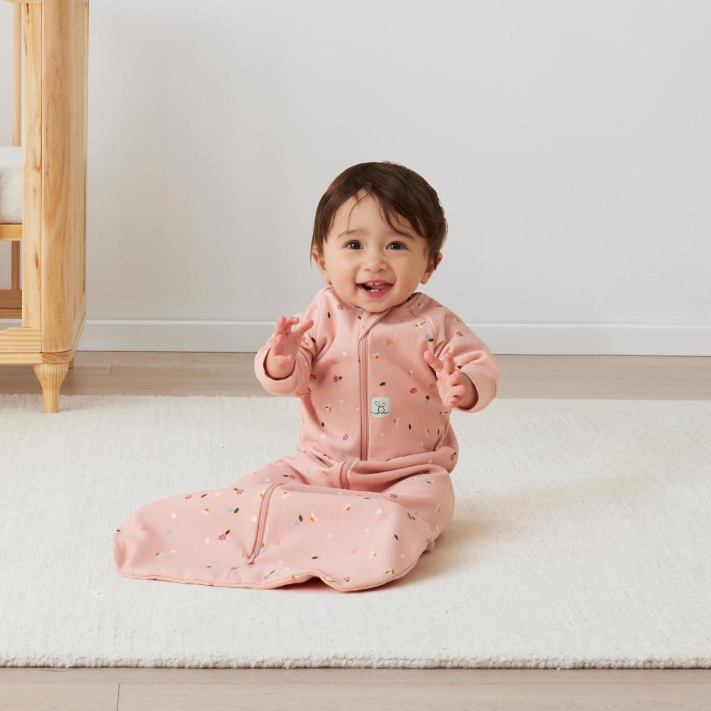 Baby in a pink sleeping bag sitting on a white rug with a wooden chair in the background