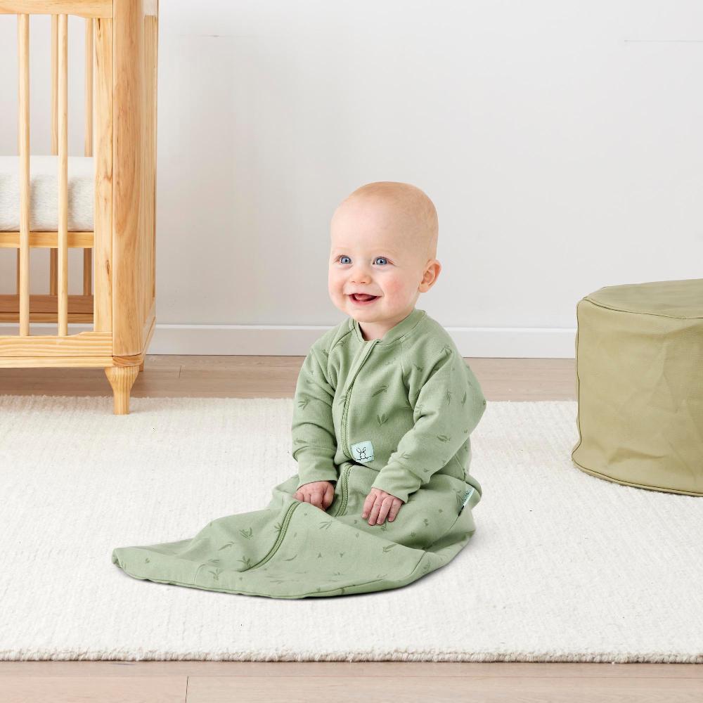 Baby in a green sleeping bag sitting on a white rug in a room with wooden furniture.