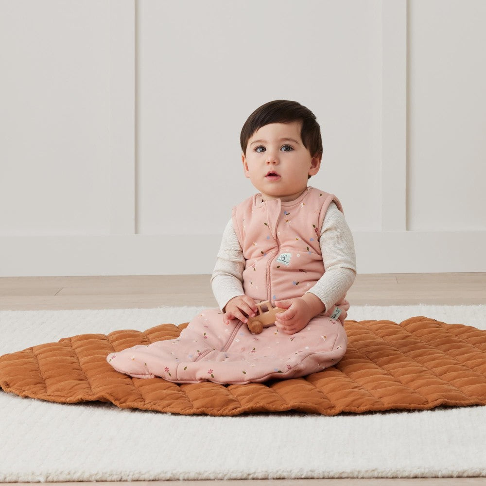 Baby sitting on a brown mat wearing a pink vest in a room with white paneled walls.
