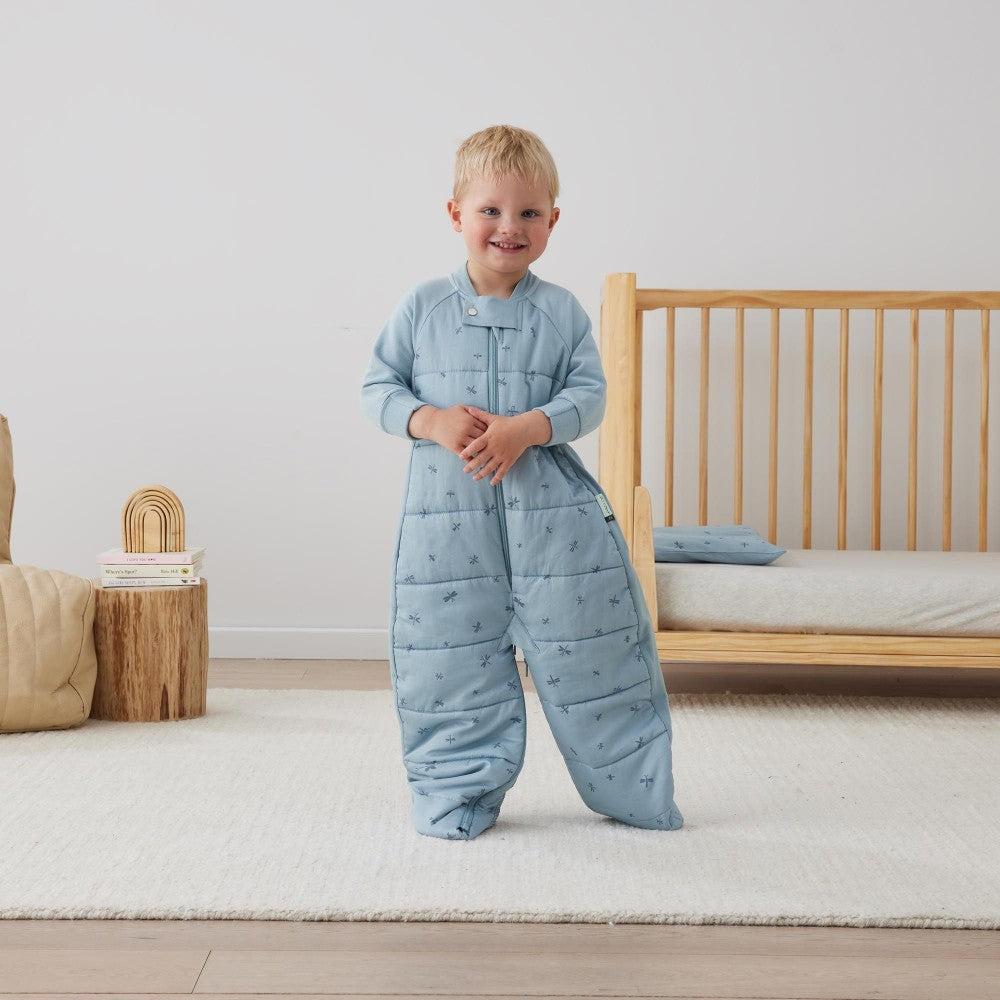 Child wearing a blue quilted outfit standing in a bedroom with a wooden crib and neutral decor.
