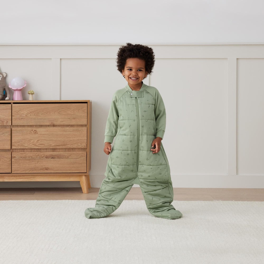 Child wearing a green quilted onesie standing in a room with wooden furniture.