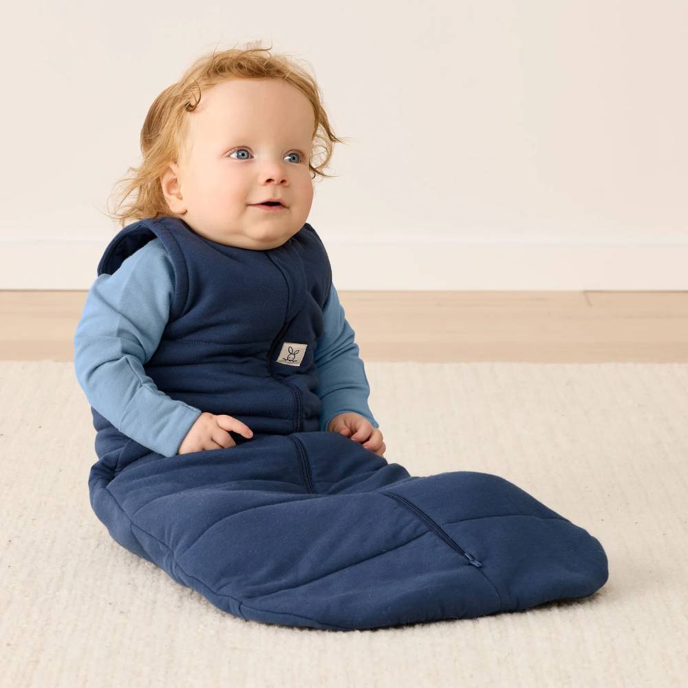 Baby wearing a blue sleeping bag sitting on a beige surface.