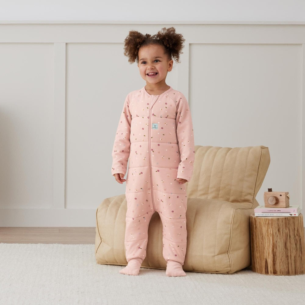 Child wearing a pink onesie with a pattern, standing in a room with a beige armchair and wooden side table.