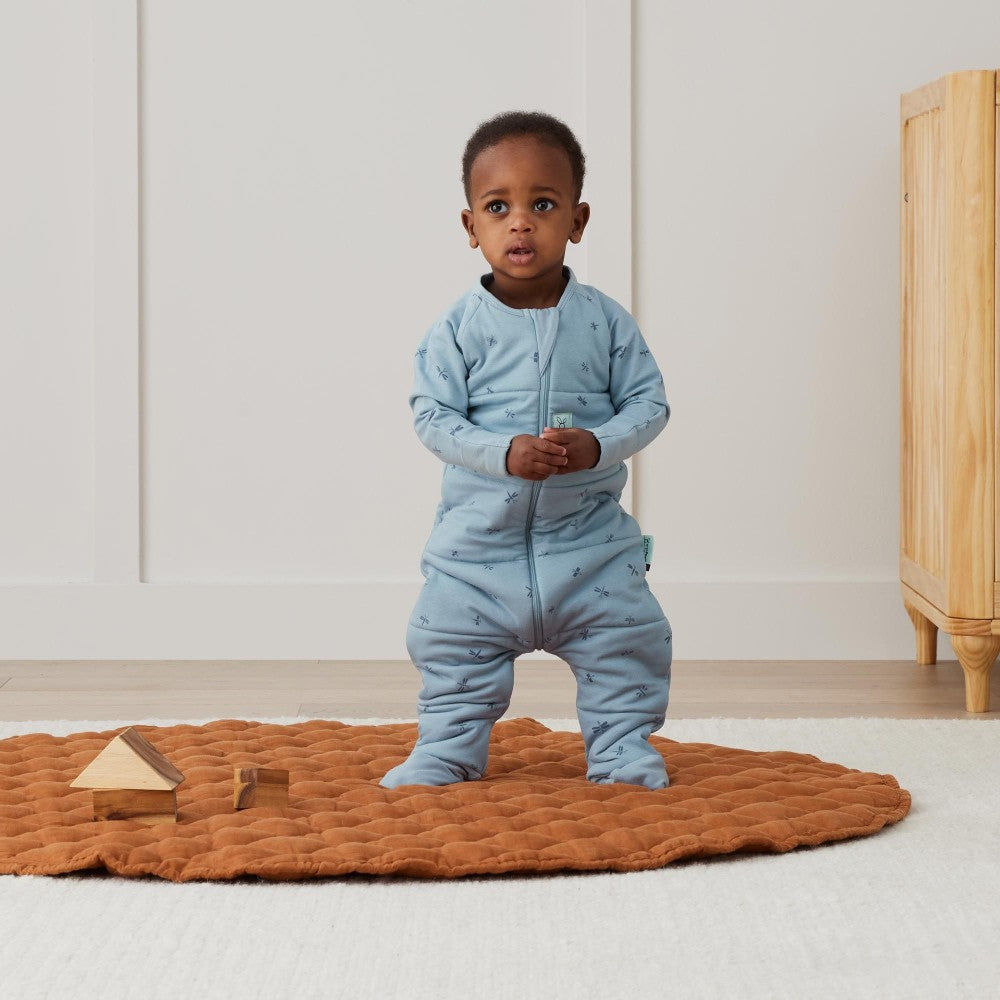 Child wearing a blue onesie standing on a brown mat with wooden toys in a room.
