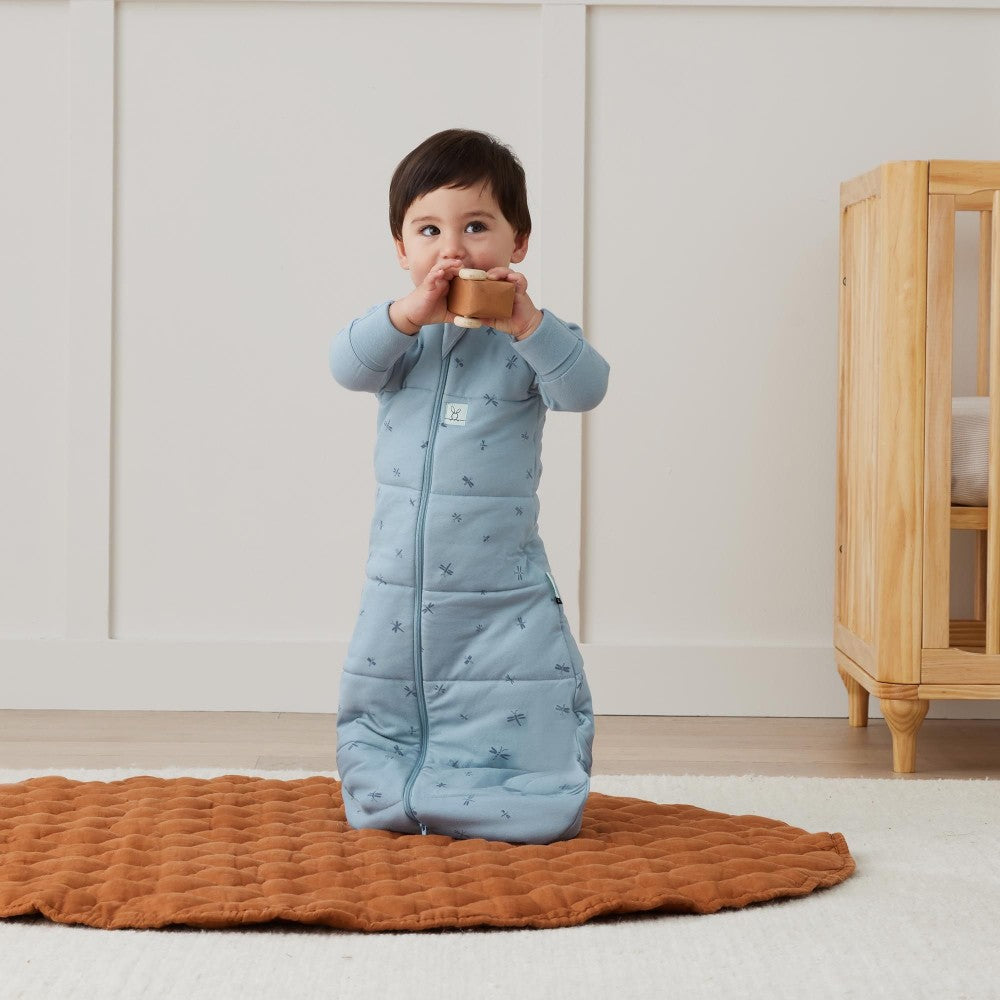 Child in a blue sleeping bag holding a wooden cup in a room with a wooden cabinet.