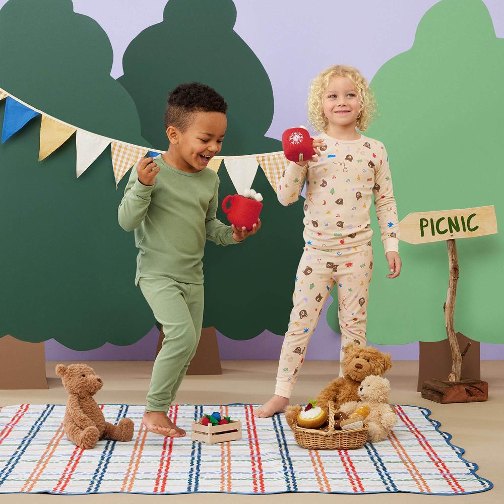 Two children in pajamas playing with toys on a picnic blanket against a forest backdrop.