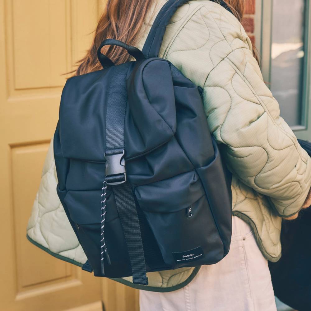 Person wearing fashionable black nappy bag backpack with a beige quilted jacket.