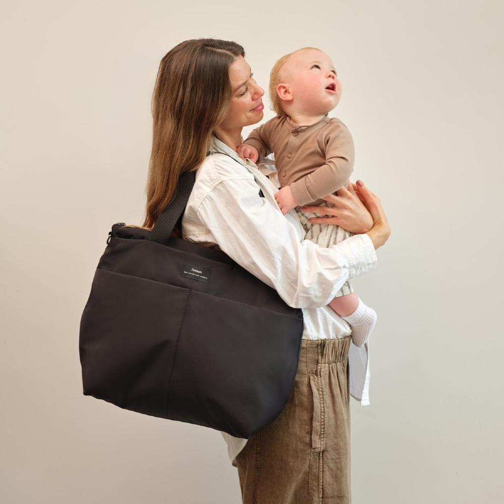 Woman holding a baby and a Carifit large fashionable black nappy bag against a plain background