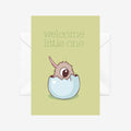 A green card with a white envelope behind it. The card has a baby kiwi hatching from an egg with the words WELCOME LITTLE ONE at the top. 