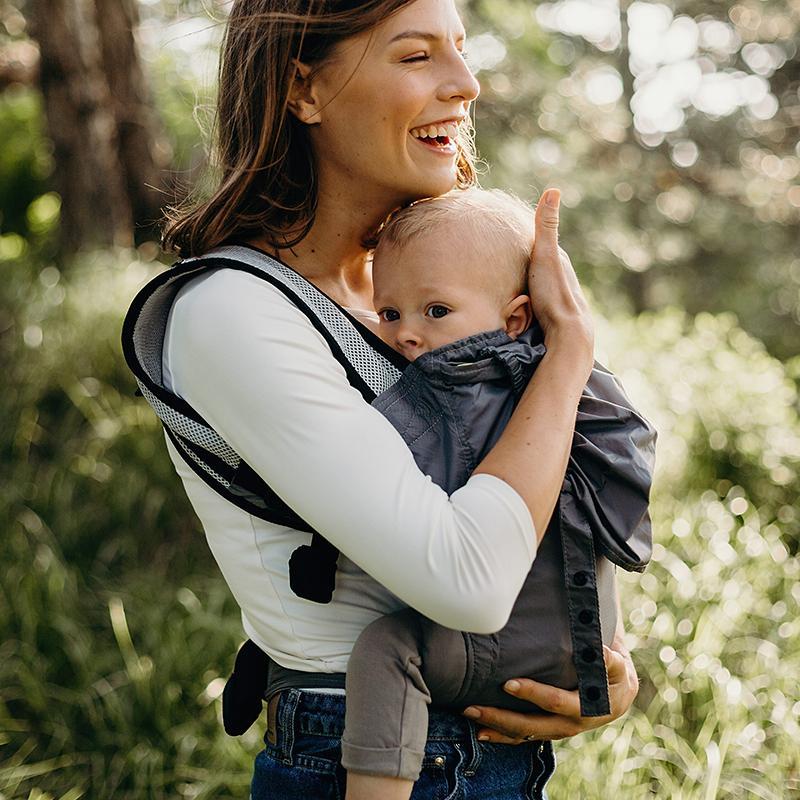 Woman carrying a baby in a compact travel carrier outdoors with greenery in the background