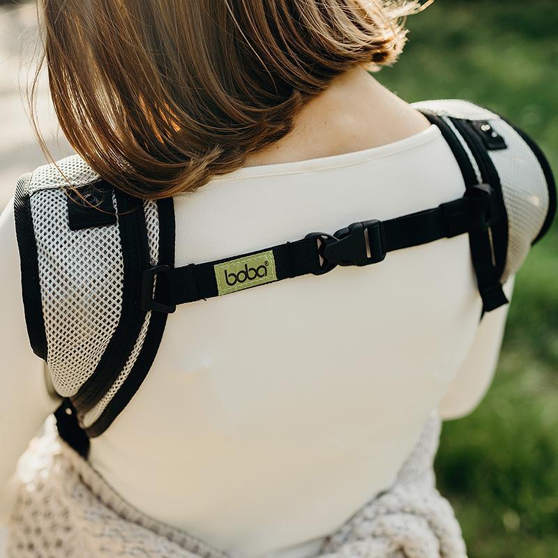 Person wearing a baby carrier with a visible brand logo, set against a blurred natural background.