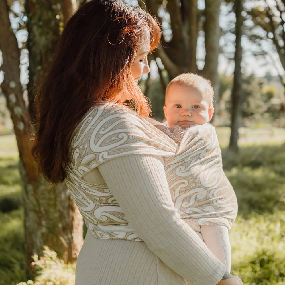 A woman outdoors carrying a baby in a cream-colored baby wrap from Hiwa with teh Okooko Pepi pattern. The background shows a soft-focus park setting with trees and grass.