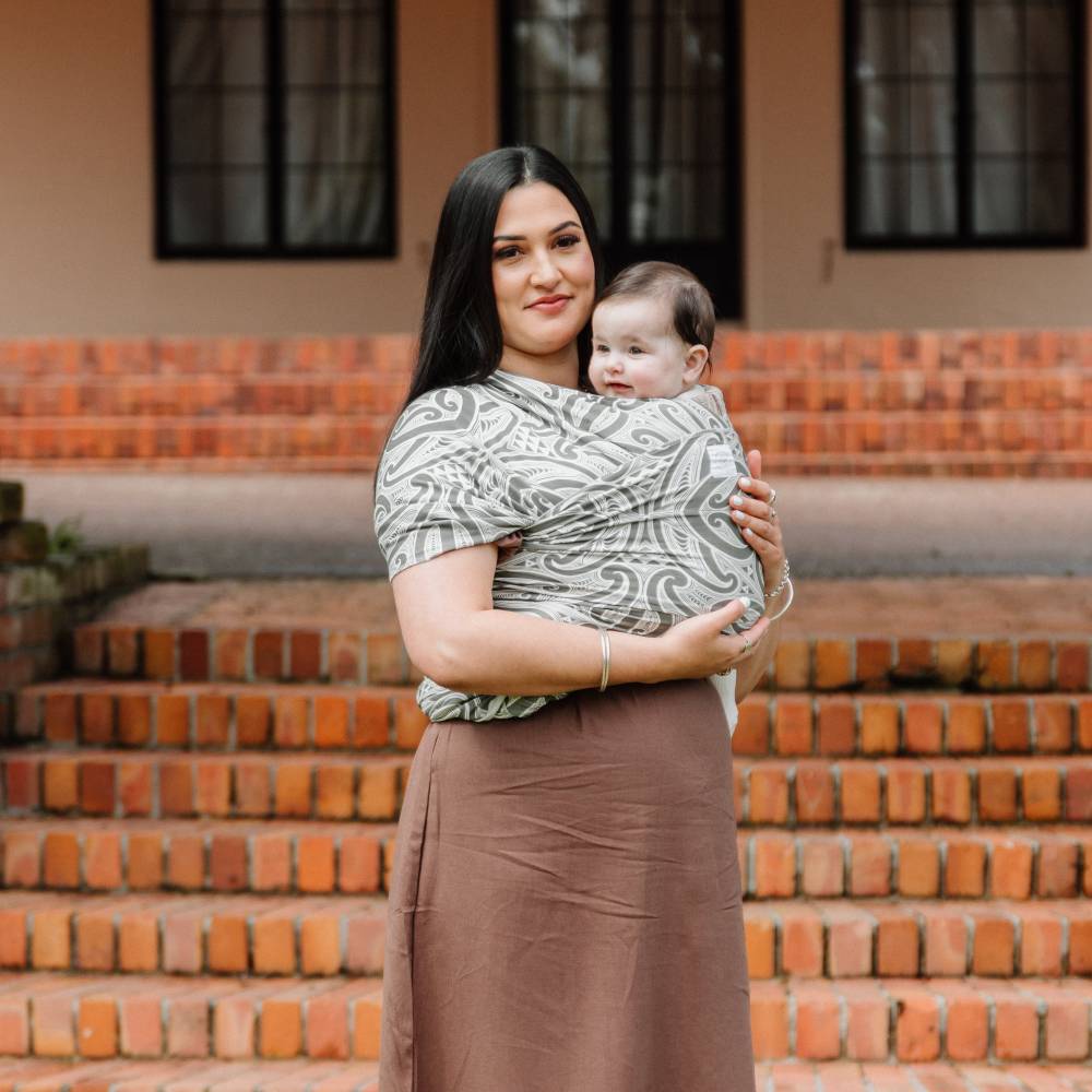A woman wearing a gray and white Hiwa Okooko Pepi patterned baby wrap carrier holds a baby in front of a brick staircase and building facade.
