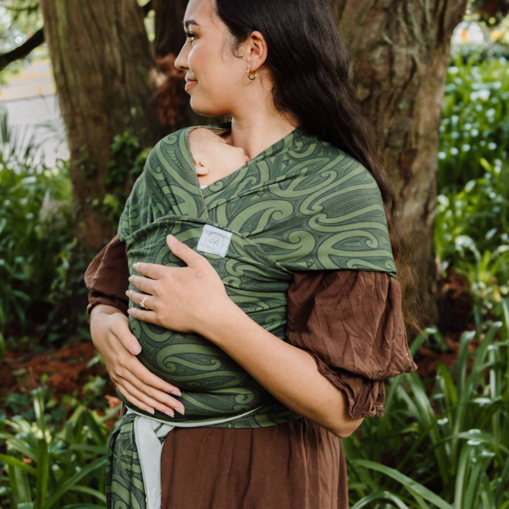 A woman outdoors wearing a baby carrier wrap in olive green with a New Zealand indigenous Maori swirling pattern and brown puff sleeves, holding a baby against her chest.