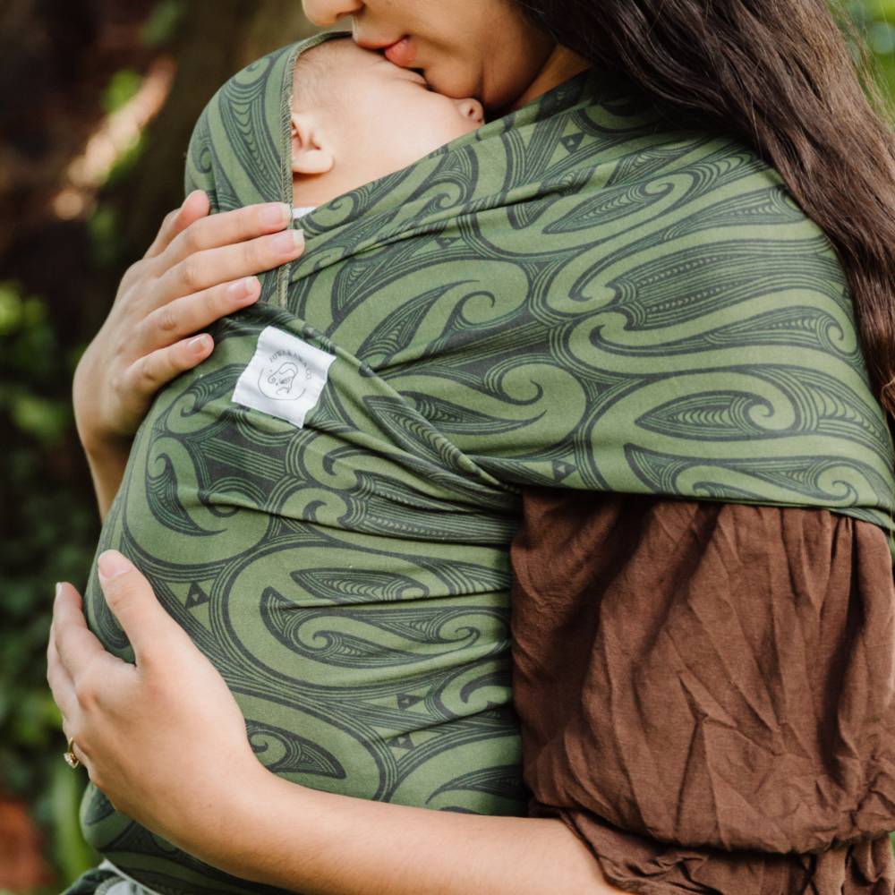 A close-up lifestyle shot of a woman wearing a Hiwa Maori print green and brown baby wrap carrier with a sleeping baby tucked inside, outdoors with a blurred natural background.