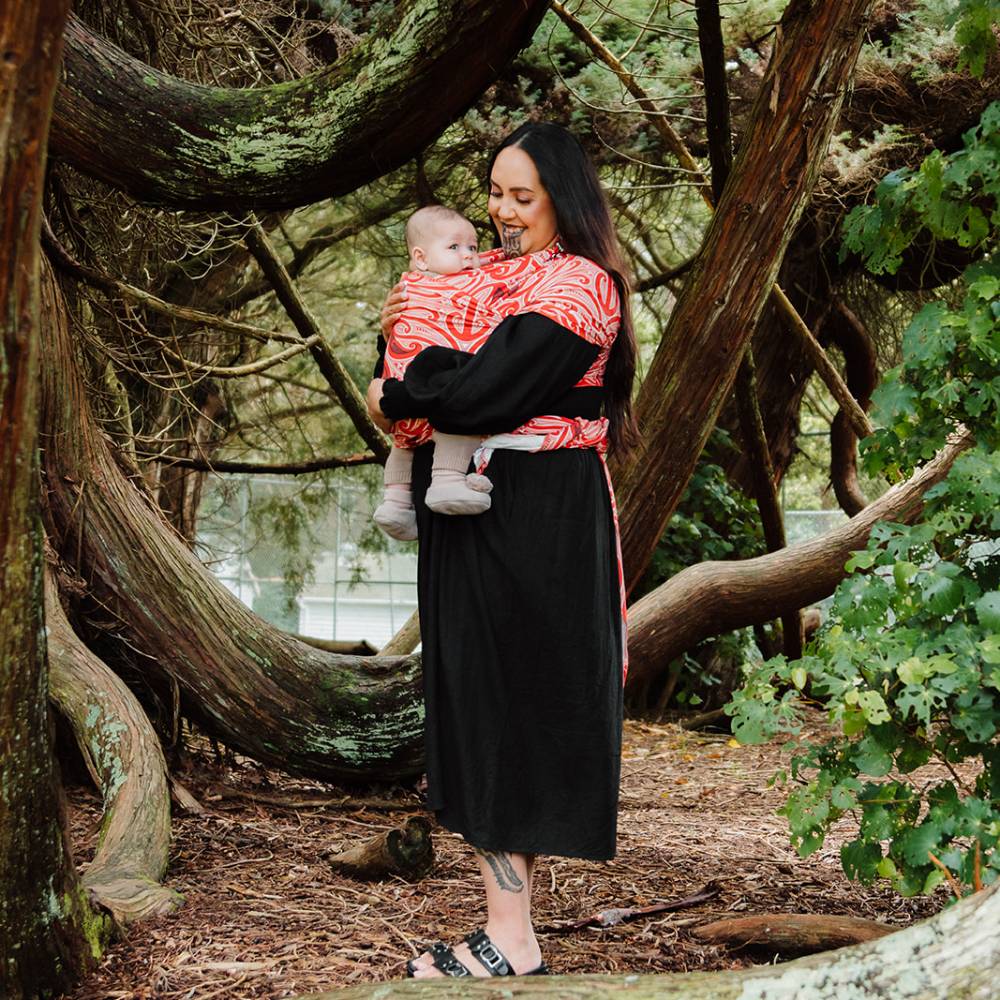 A woman carrying a baby outdoors in a forest, wearing a Maori patterned wrap-style top with a red and black pattern and a black skirt.