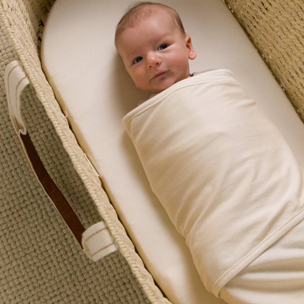Newborn baby wrapped in a white miracle blanket swaddle lying in a beige crib with cream handles