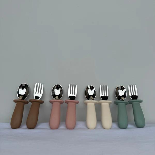 Six pairs of baby utensils with matte-colored handles are lined up against a neutral gray background. The handles come in brown, dusty pink, cream, and sage green, paired with silver metal spoons and forks.
