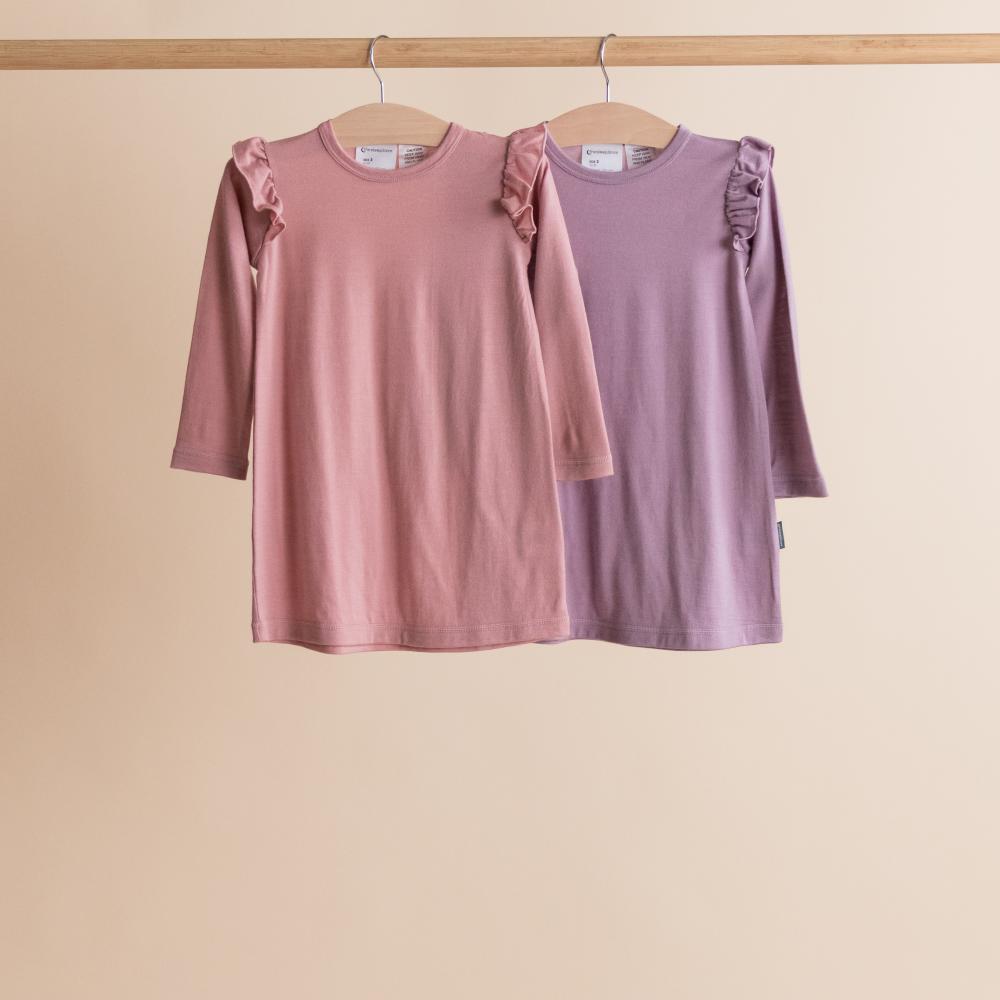 Two children's nighties with ruffled shoulders hanging on a wooden rod against a beige background.
