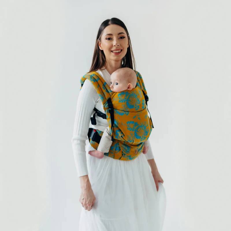 A woman holding her baby in a baby carrier. The carrier is a mustard colour with a teal green floral pattern. 