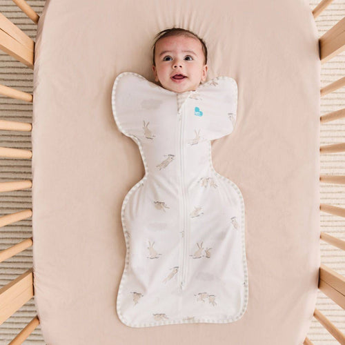 Baby lying in a crib with a white sleep swaddle featuring a bunnies prints.