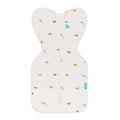 An off-white baby sleep swaddle with a front zipper, decorated with a small pattern of colorful dinosaurs and a small blue heart logo on the chest.