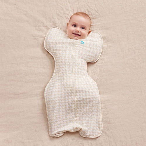 A baby lying on a beige blanket, wrapped in a baby swaddle with a small blue tag near the neck.