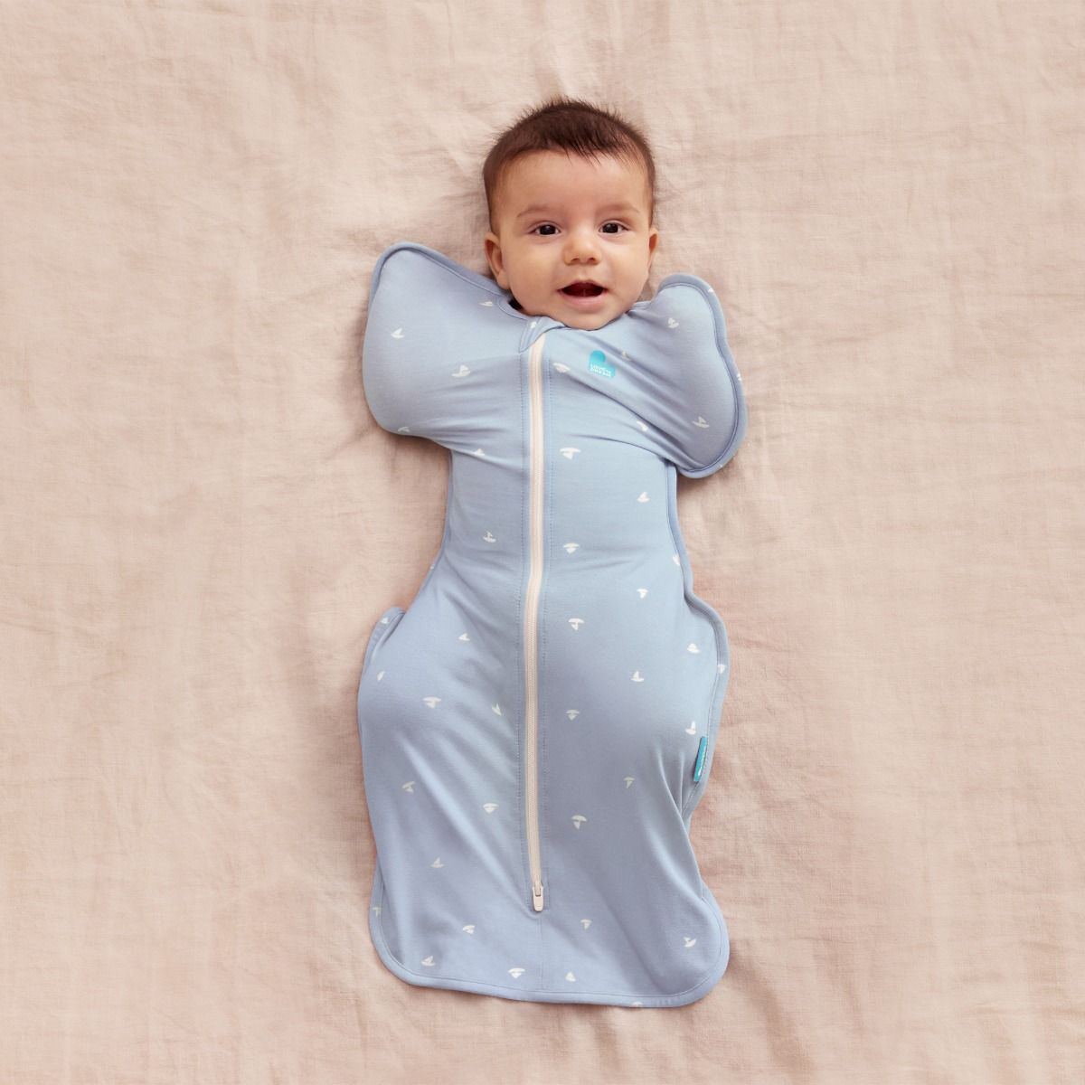 A baby lying down wearing a blue fitted swaddle with an all over boats design. 