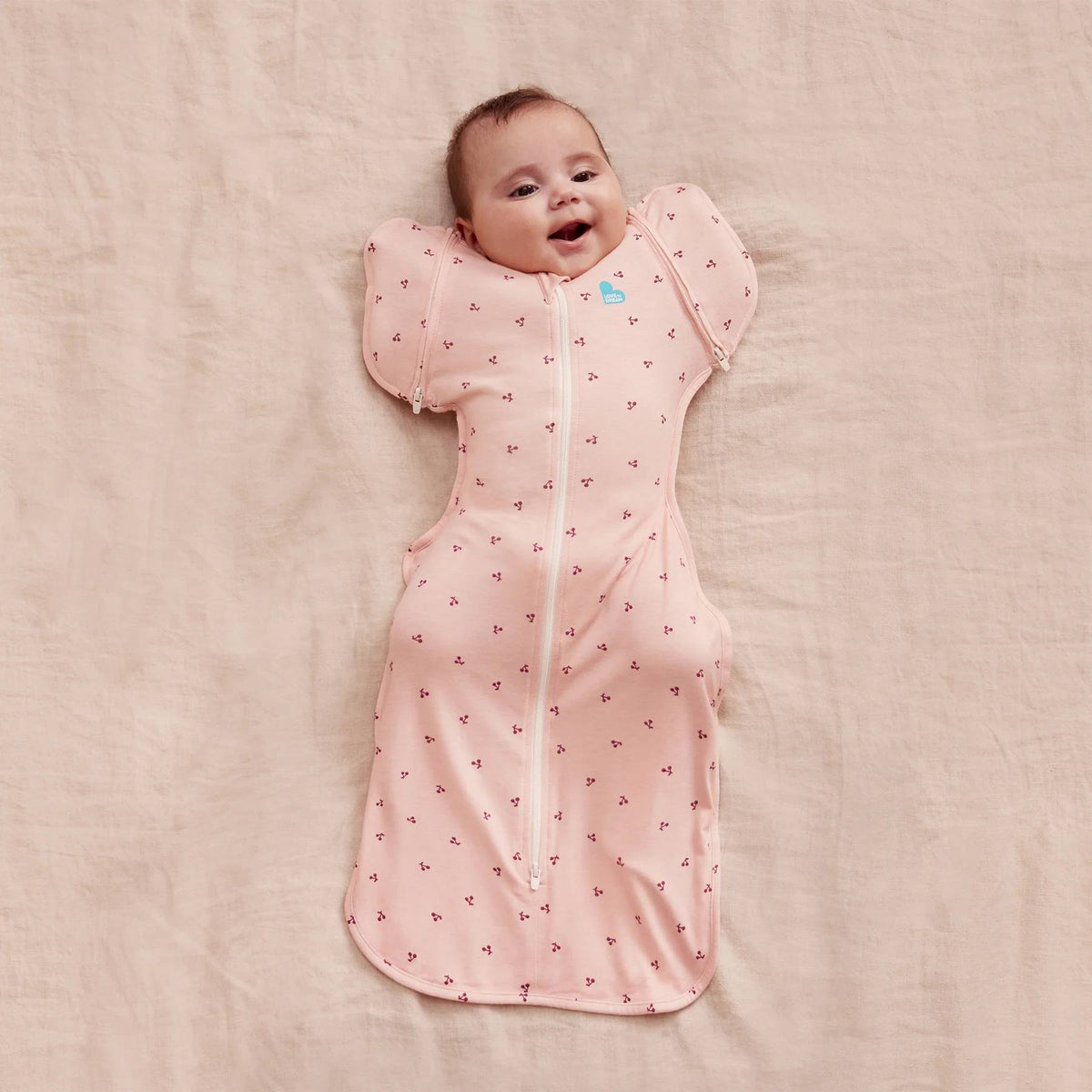 A baby lying down wearing a pink fitted swaddle with an all over cherries print. 