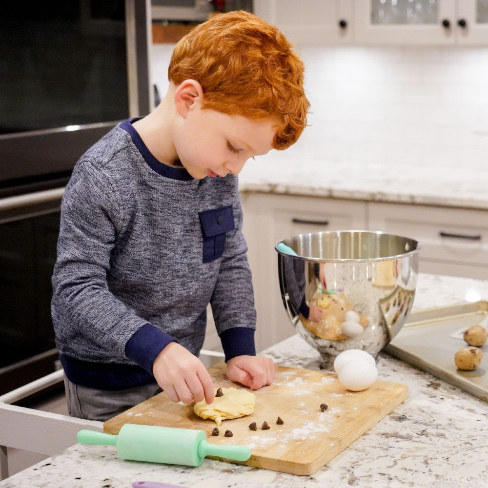 Child in a kitchen preparing food on a cutting board with a bowl and rolling pin in the background.