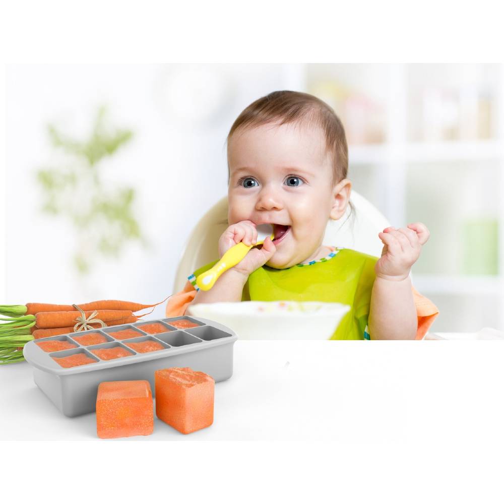 Baby eating with a spoon next to a tray of food and carrots