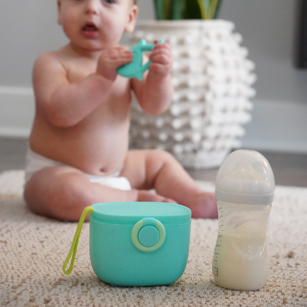 Baby sitting on a carpeted floor with a teething toy, cup, and bottle in front of them.
