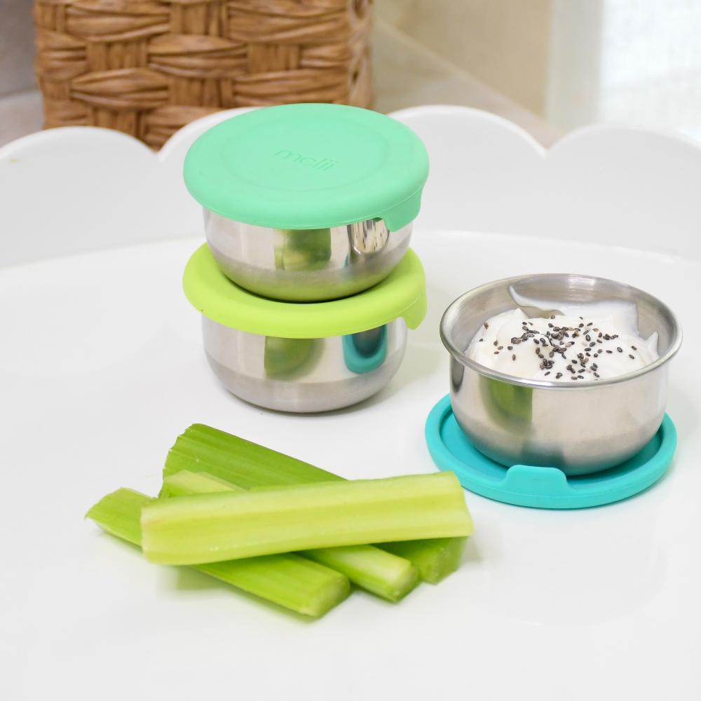 Two stainless steel containers with green and blue lids on a white surface with celery sticks.