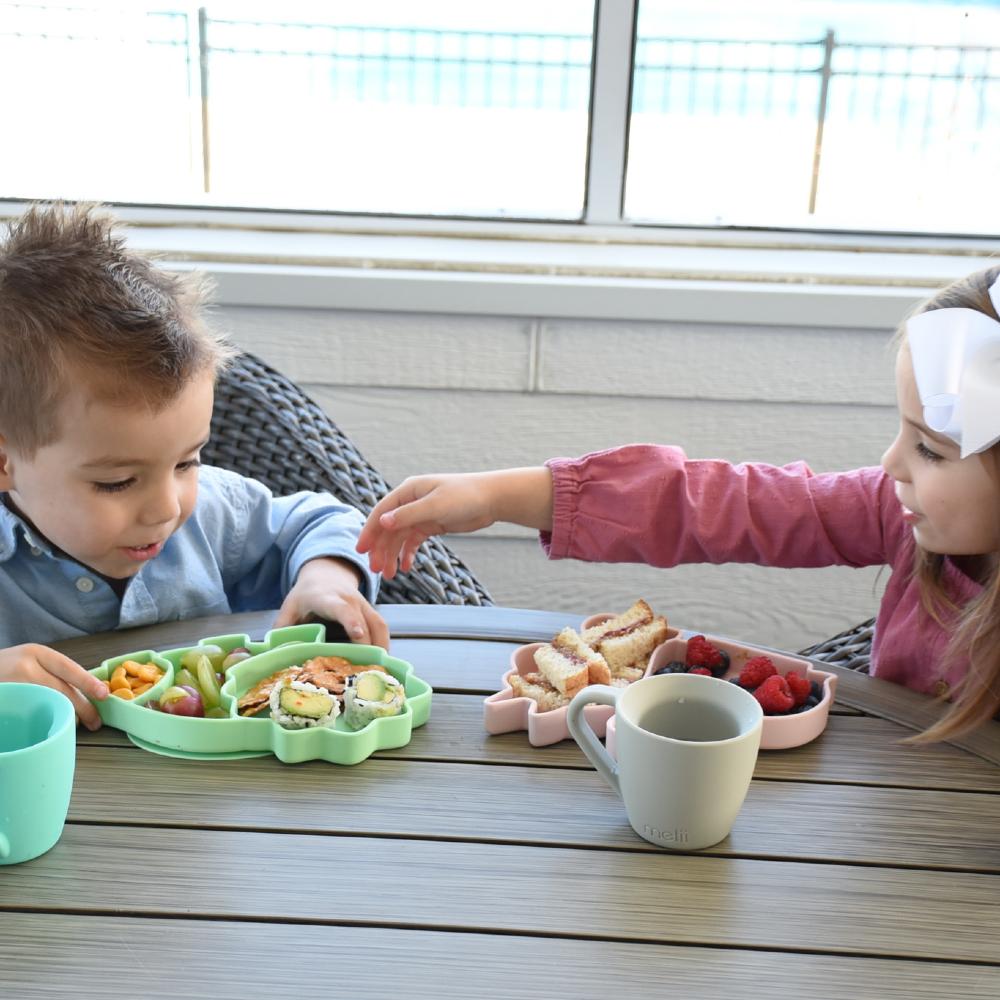 Two children sitting at a table with food and drinks, with melii divided animal shaped plates