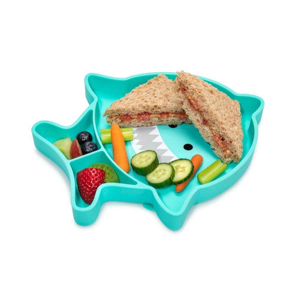 Shark-shaped silicone divided plate with a sandwich, vegetables, and fruit on a white background