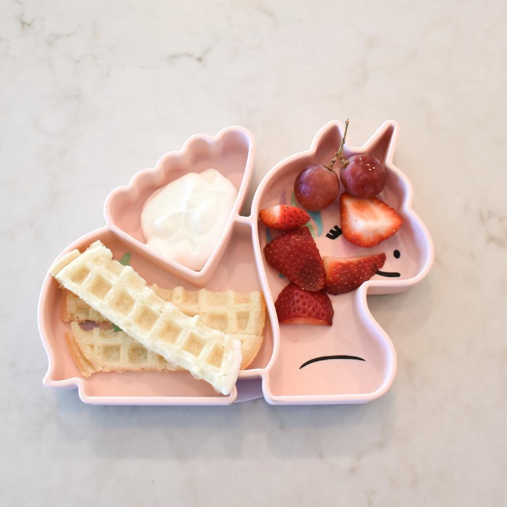 Pink children's lunchbox with compartments containing waffles, strawberries, and grapes on a marble surface.