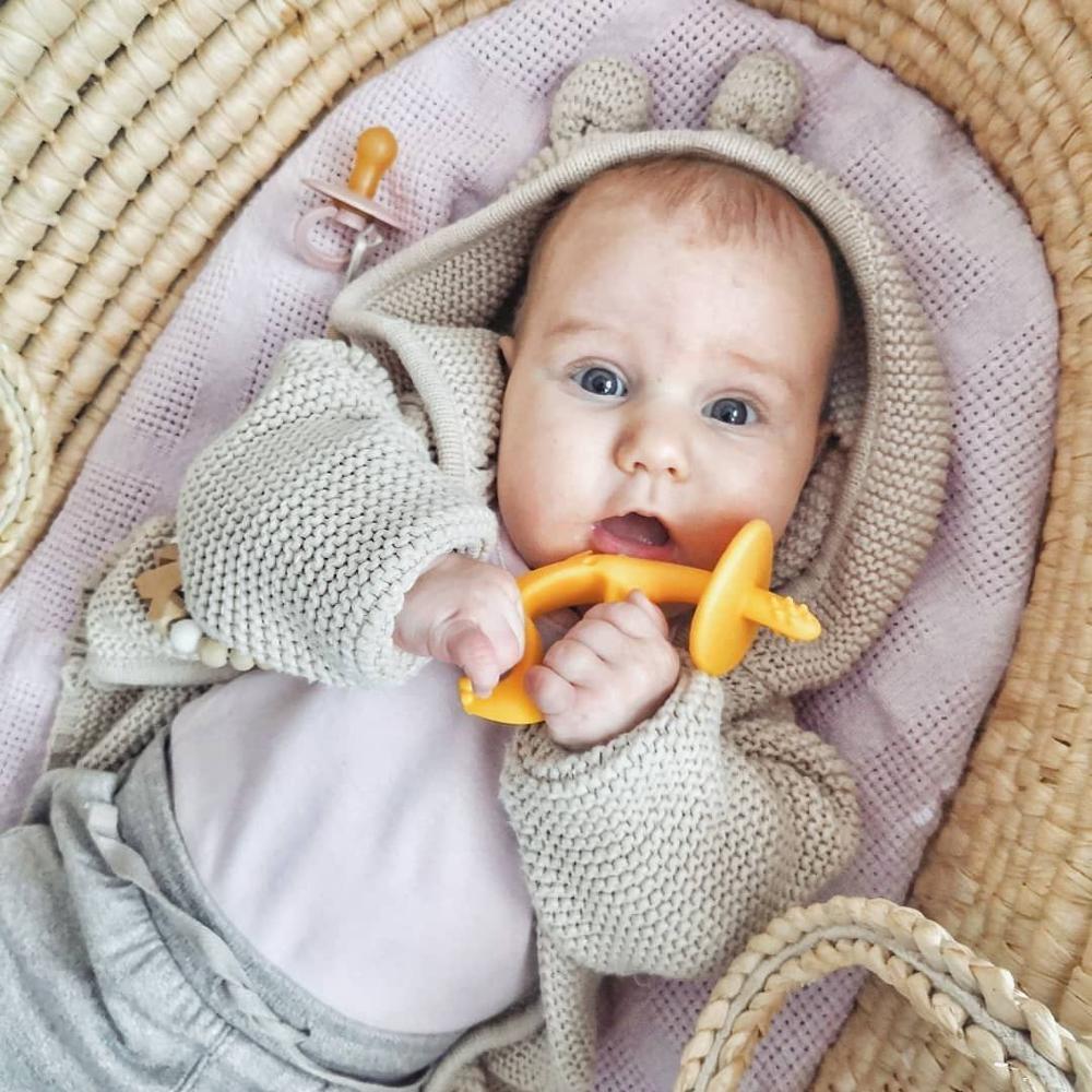 A baby wearing a beige knitted hooded outfit with bear ears, lying in a woven basket on a pink blanket and holding a yellow teether.